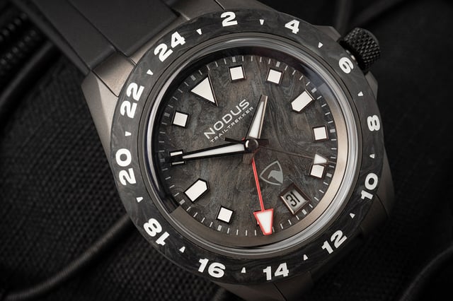 Nodus x Raven TrailTrekker Carbon dial detail — marbled forged carbon with polished gunmetal indices, red-tipped GMT hand, Raven shield logo above date at 6