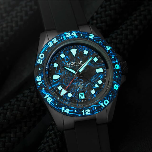 Nodus x Raven TrailTrekker Carbon lume shot — forged carbon bezel and dial glowing blue in darkness, entire marbled grain illuminated with Super-LumiNova